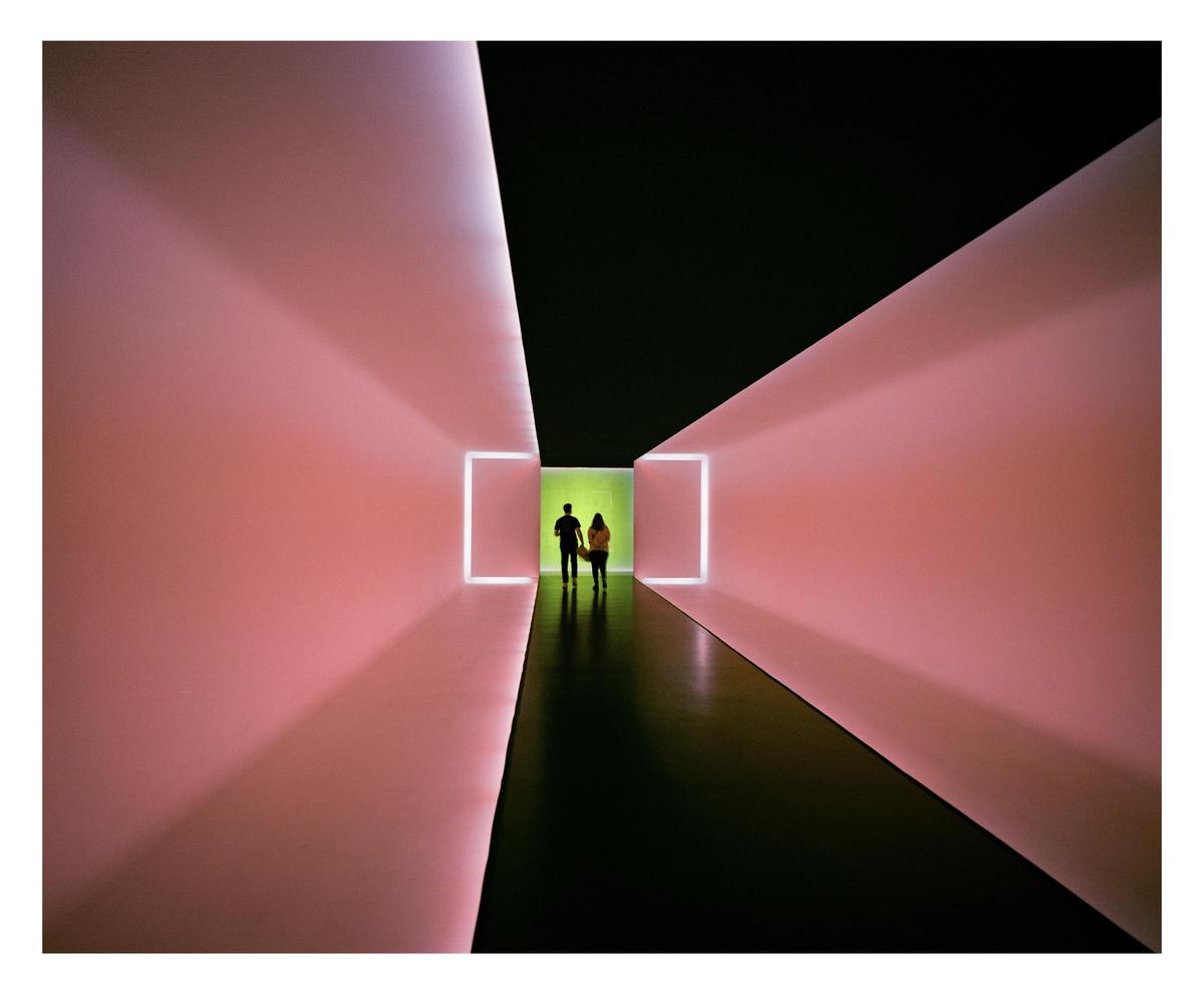 Two people stand in a brightly lit hallway with pink walls and a green light at the end.