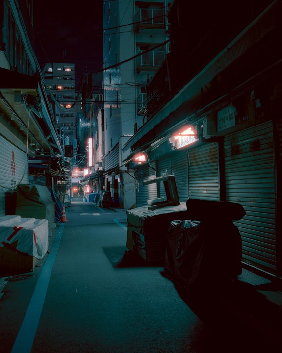 A narrow alleyway is lined with closed shops and buildings under a dark night sky.