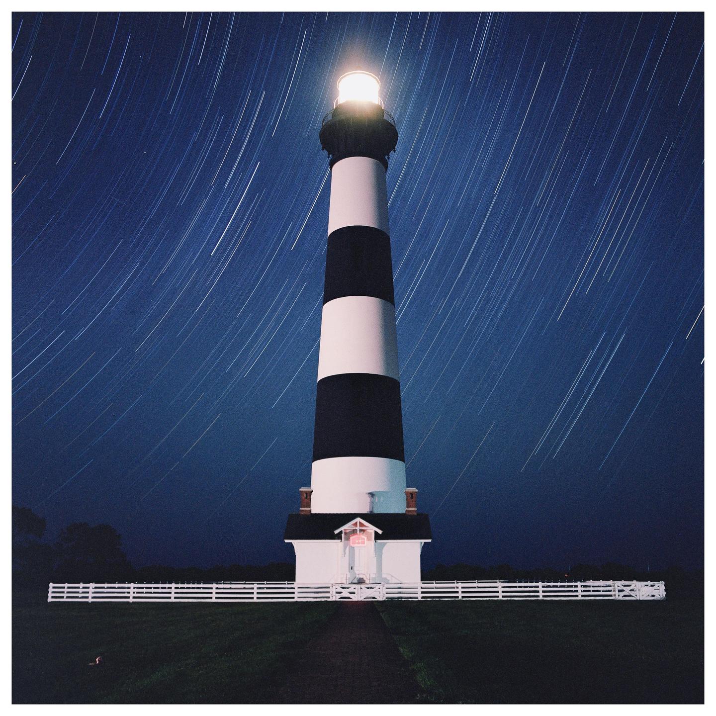 A black and white striped lighthouse stands tall against a dark blue night sky with star trails.