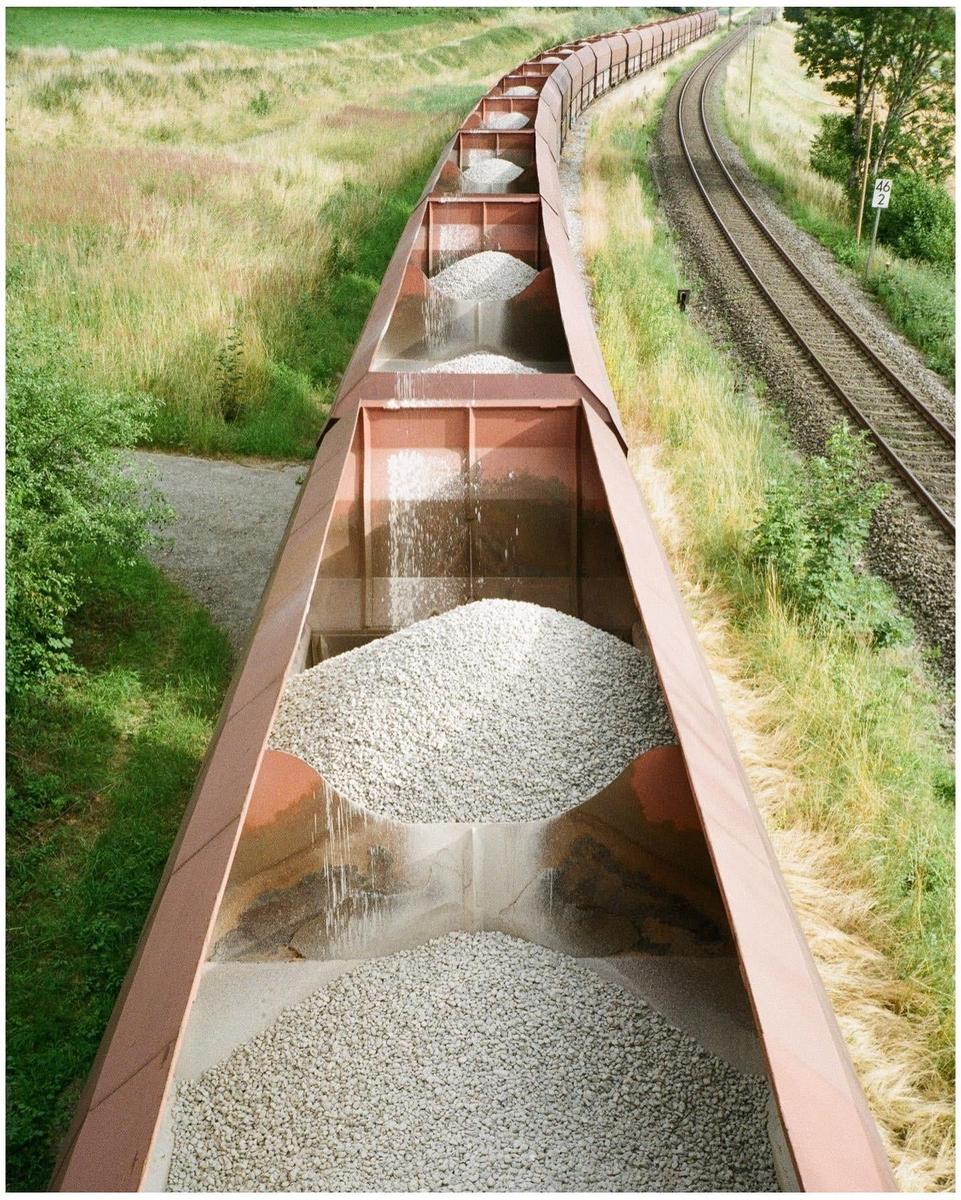 A long train with open wagons filled with gravel is moving along a railway track next to a grassy field.