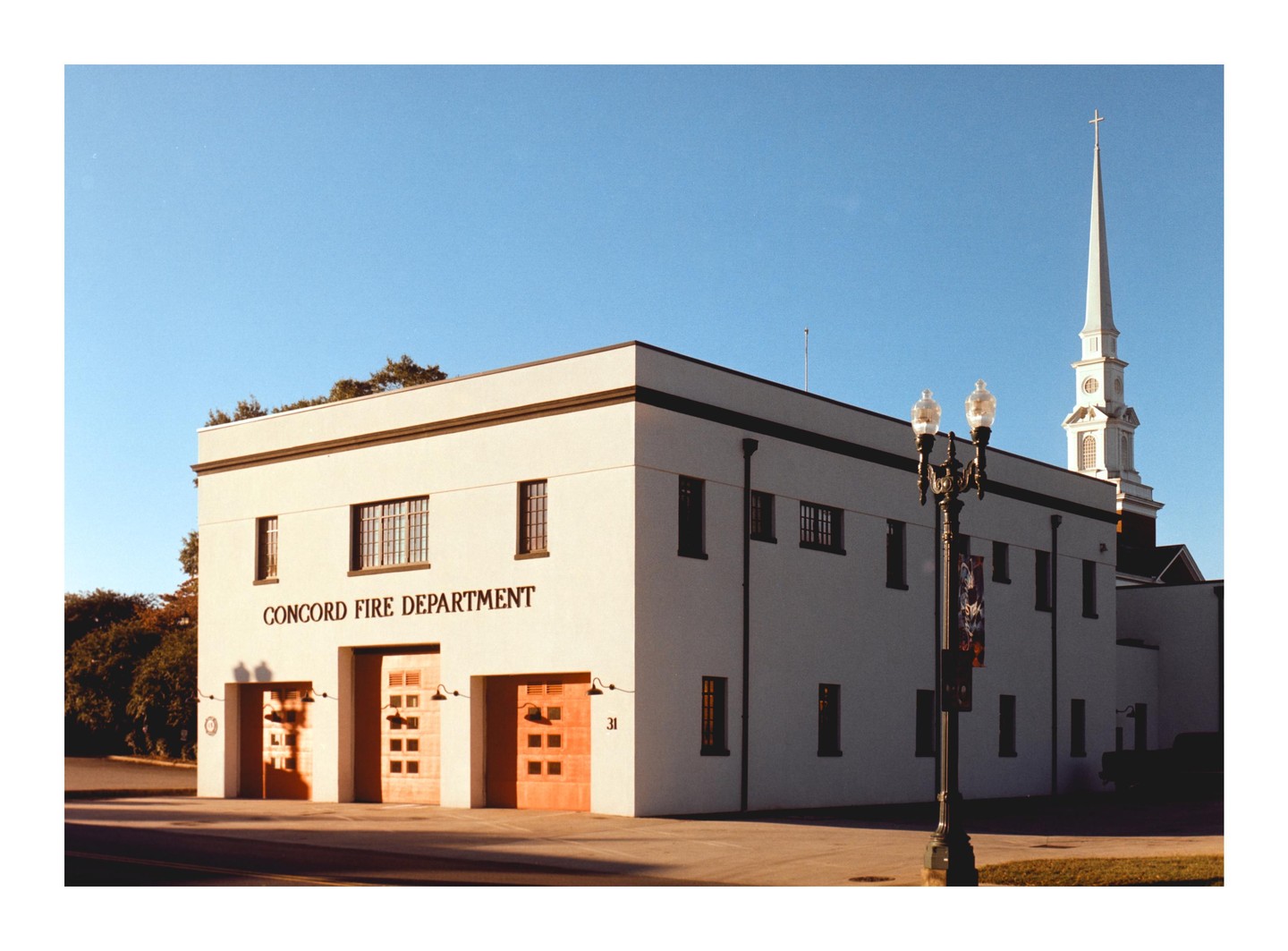 The Concord Fire Department building with a church steeple in the background is illuminated by morning light.