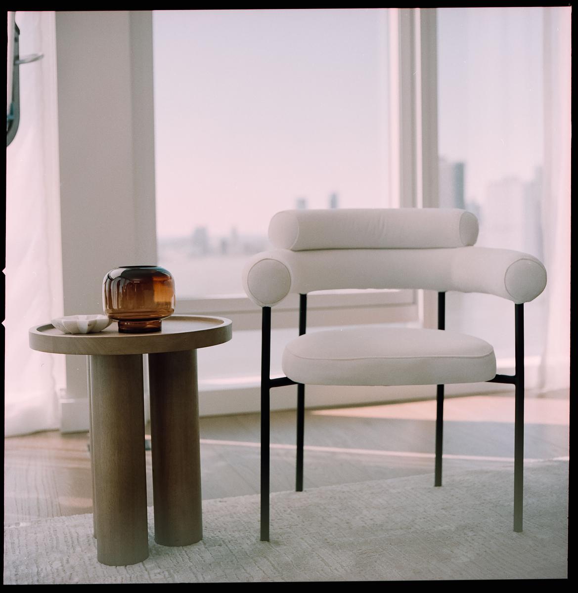 A modern white chair and a round side table with a brown vase are positioned in front of a large window overlooking a city.