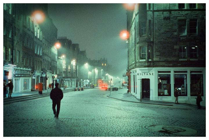 A man walks down a cobblestone street at night with buildings and streetlights on either side.