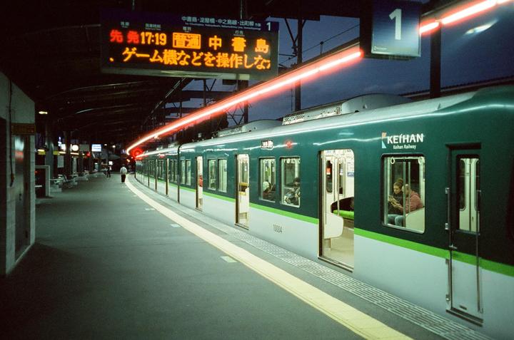 A green and white train is stopped at a station platform with a red light trail streaking across the top of the image.