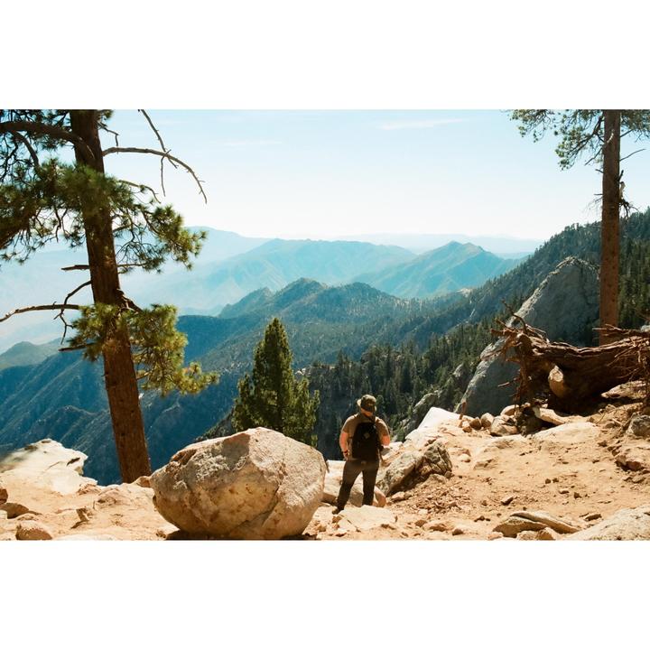 A man stands on a rocky mountain path looking out at a vast mountain range.