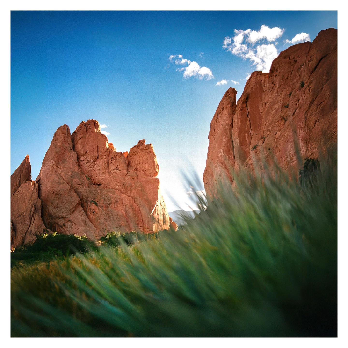 Sunlight shines between two large red rock formations with green grass in the foreground.