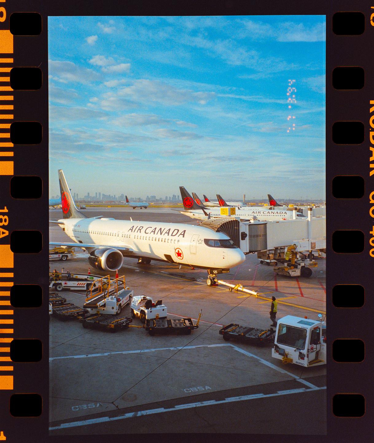 An Air Canada airplane is parked at an airport gate with other airplanes and a city skyline in the distance.