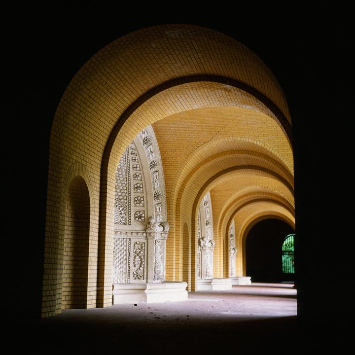 A long corridor with brick arches and decorative stone columns is illuminated by light from the right.