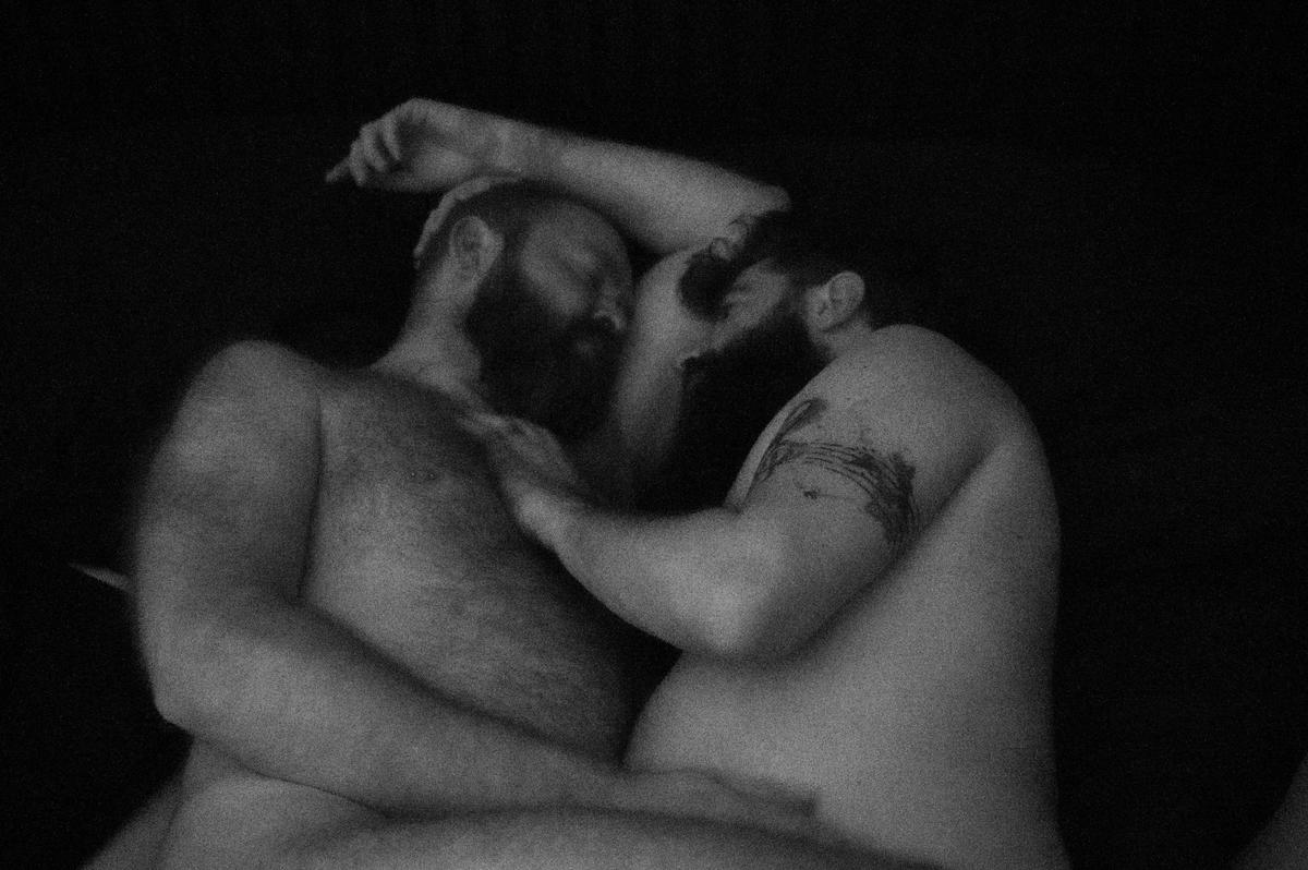 Two shirtless men with beards are embracing each other in a dark setting.