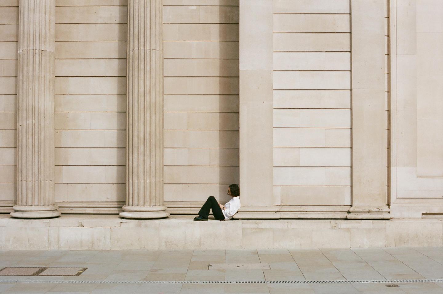 A man sits on a ledge in front of a large stone building with columns.