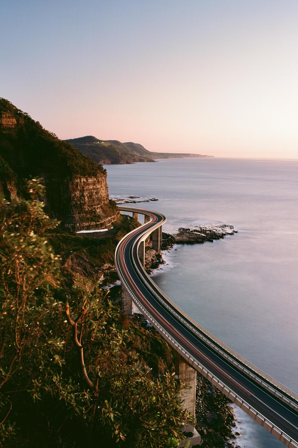 A long exposure shot captures the light trails of vehicles on the Sea Cliff Bridge as it curves along the coastline at dusk.