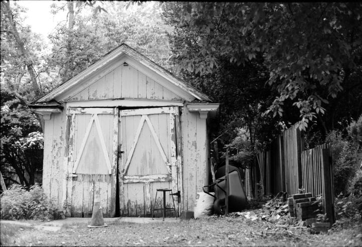 A white shed with peeling paint and two large doors stands next to a wooden fence, a wheelbarrow, and various garden items.
