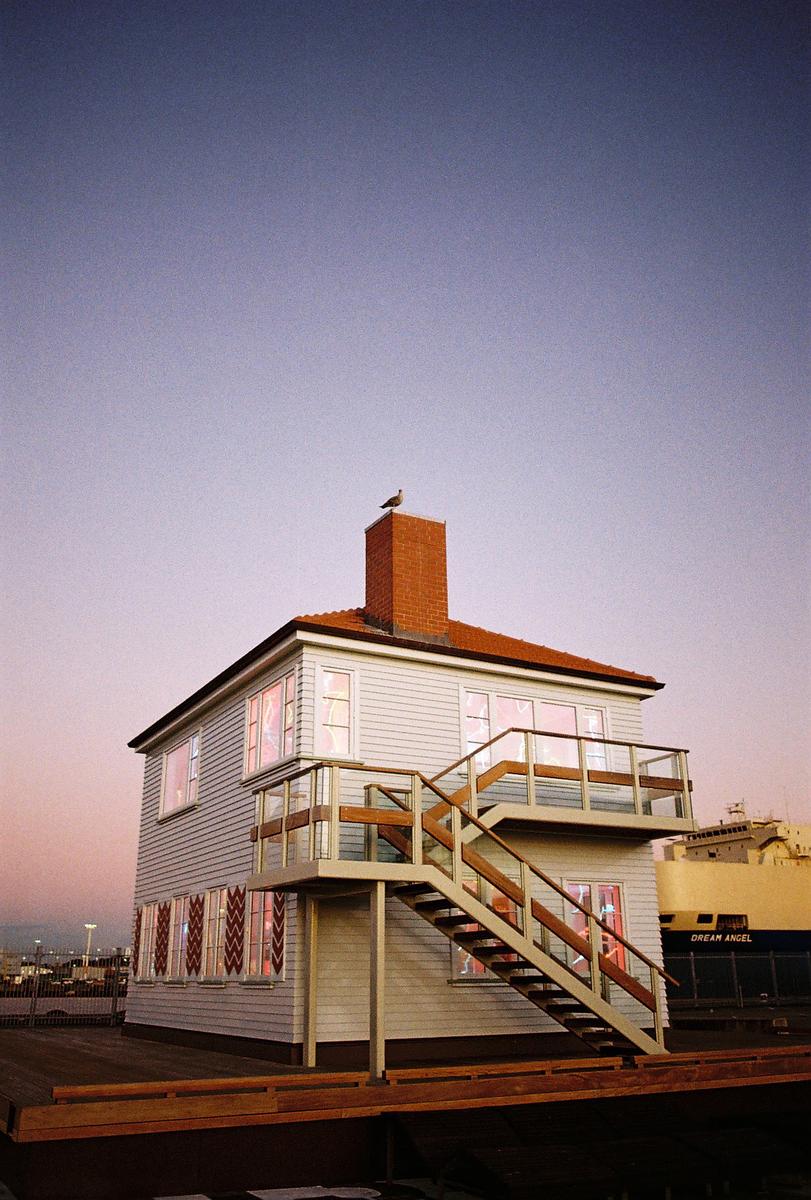 A bird sits atop a chimney on a two-story house with an external staircase, next to a large ship.