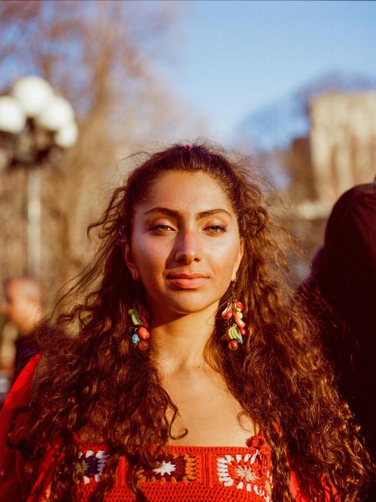 A woman with long, curly hair and colorful earrings looks directly at the viewer.
