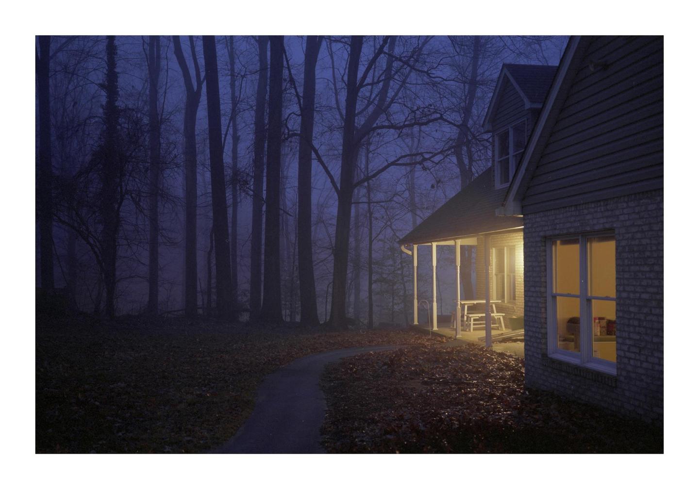 A winding path leads to a house with a lit porch and window, surrounded by a dark, hazy forest at night.