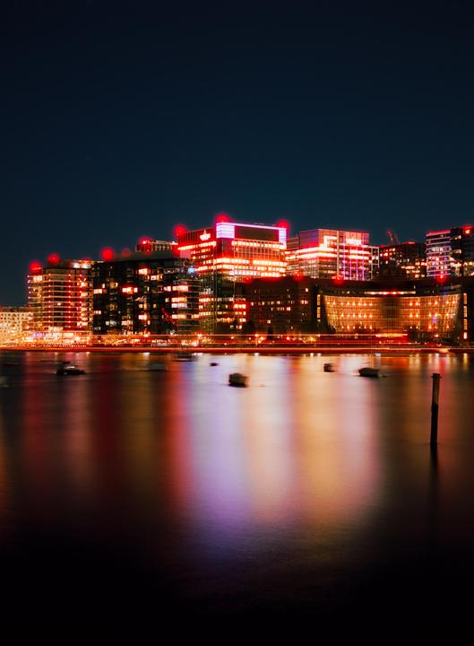 A long exposure shot of the Boston waterfront at night shows buildings lit up with red and orange lights reflecting on the water.