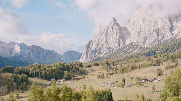 A small village with houses and trees sits in a valley surrounded by large mountains under a cloudy sky.