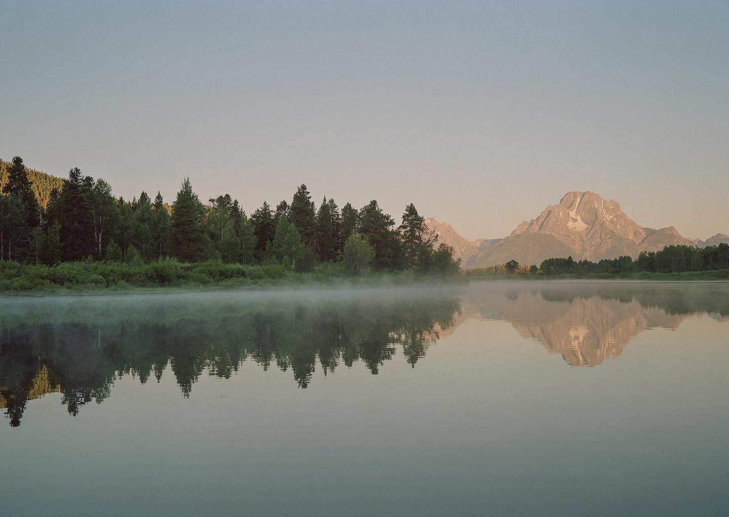 A misty lake reflects a forest and mountain under a clear sky.