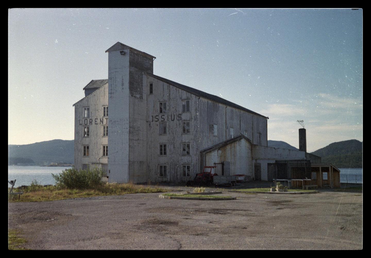 A large white building with the words "LORENT" and "ISSIUS" painted on its side stands next to a body of water with mountains in the background.