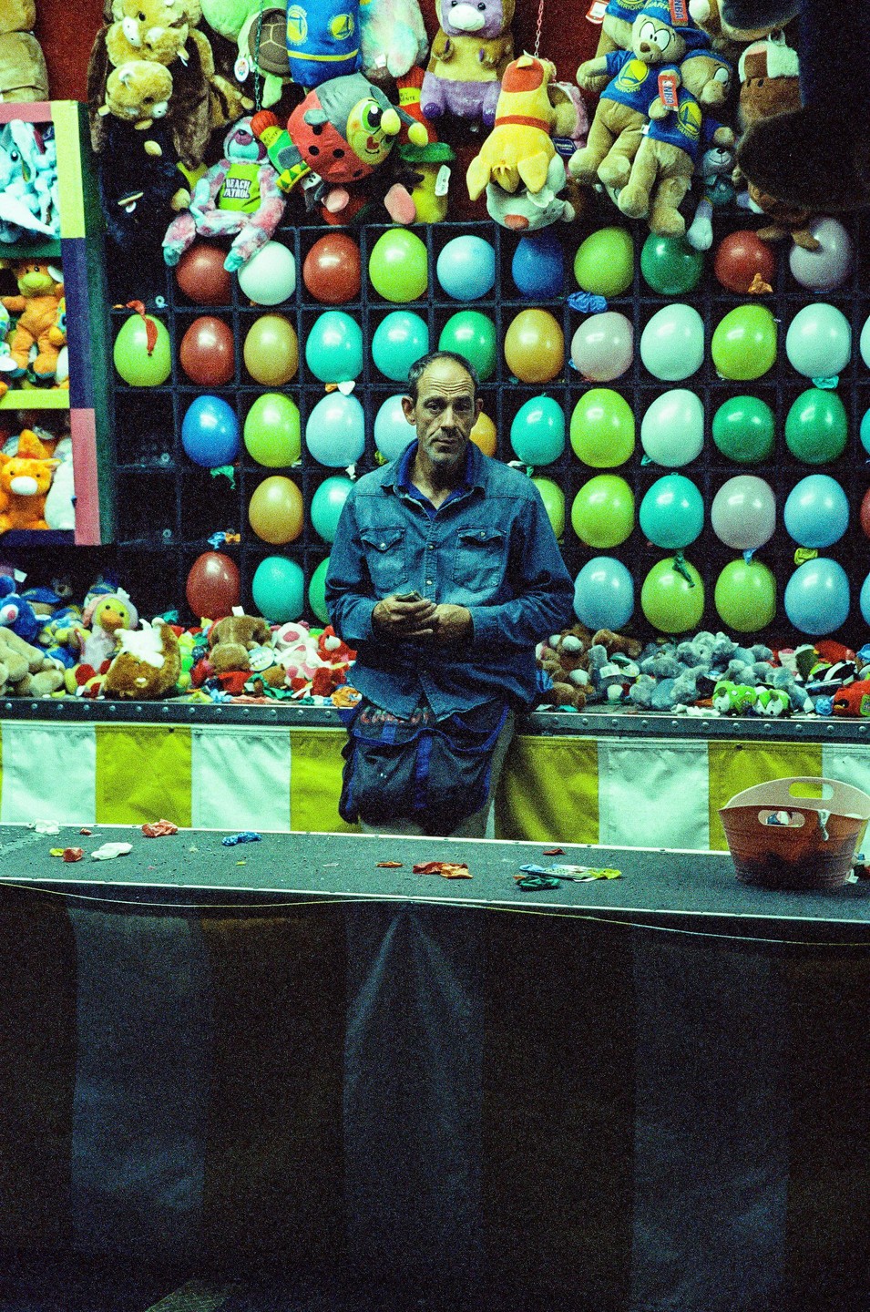 A man stands in front of a carnival game booth filled with balloons and stuffed animals.