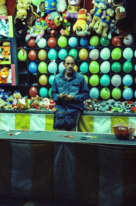 A man stands in front of a carnival game booth filled with balloons and stuffed animals.