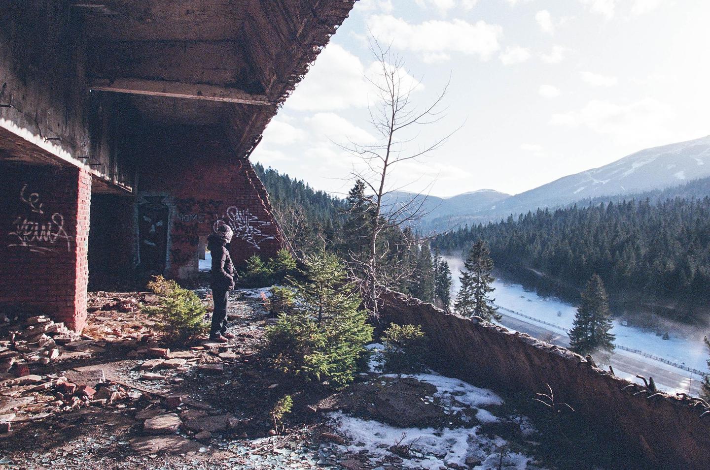 A person stands in the ruins of a building overlooking a snowy valley with a road and mountains.