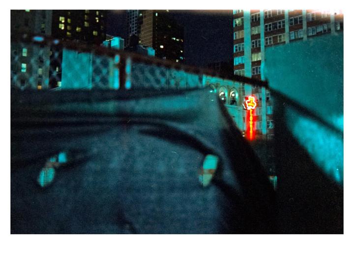 A blurry foreground frames a view of Times Square at night, with a red neon sign visible among the buildings.