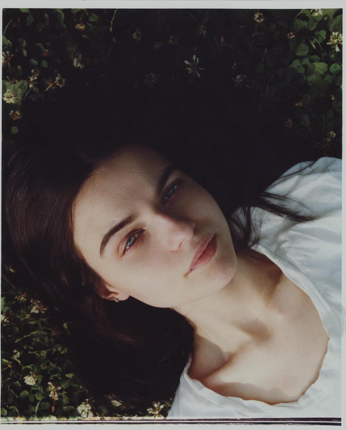A woman with dark hair and a white shirt is lying on the ground surrounded by green foliage and small white flowers.