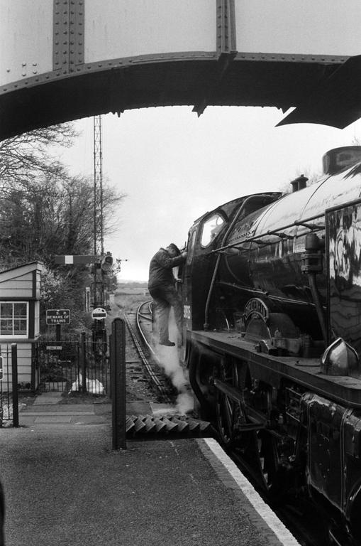 A man climbs into the cab of a steam train at a station next to a signal box.