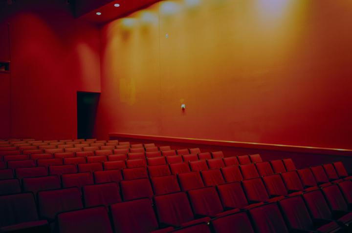 An empty auditorium with rows of red seats faces a red and orange wall with a dark doorway.