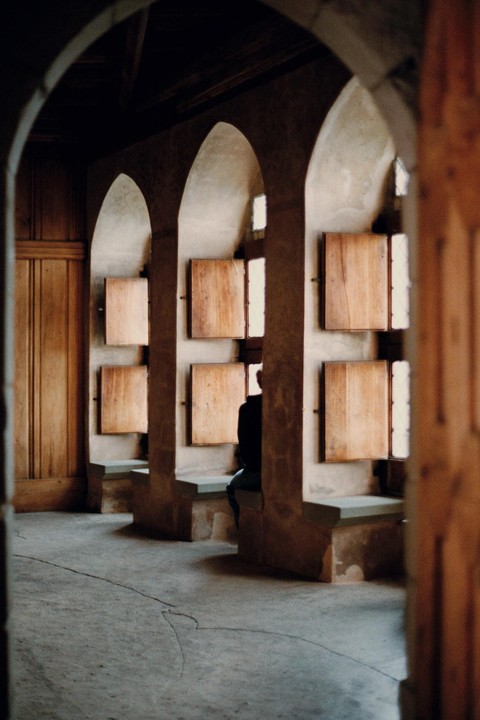 A person sits on a stone bench looking out a window with wooden shutters in a room with arched doorways.