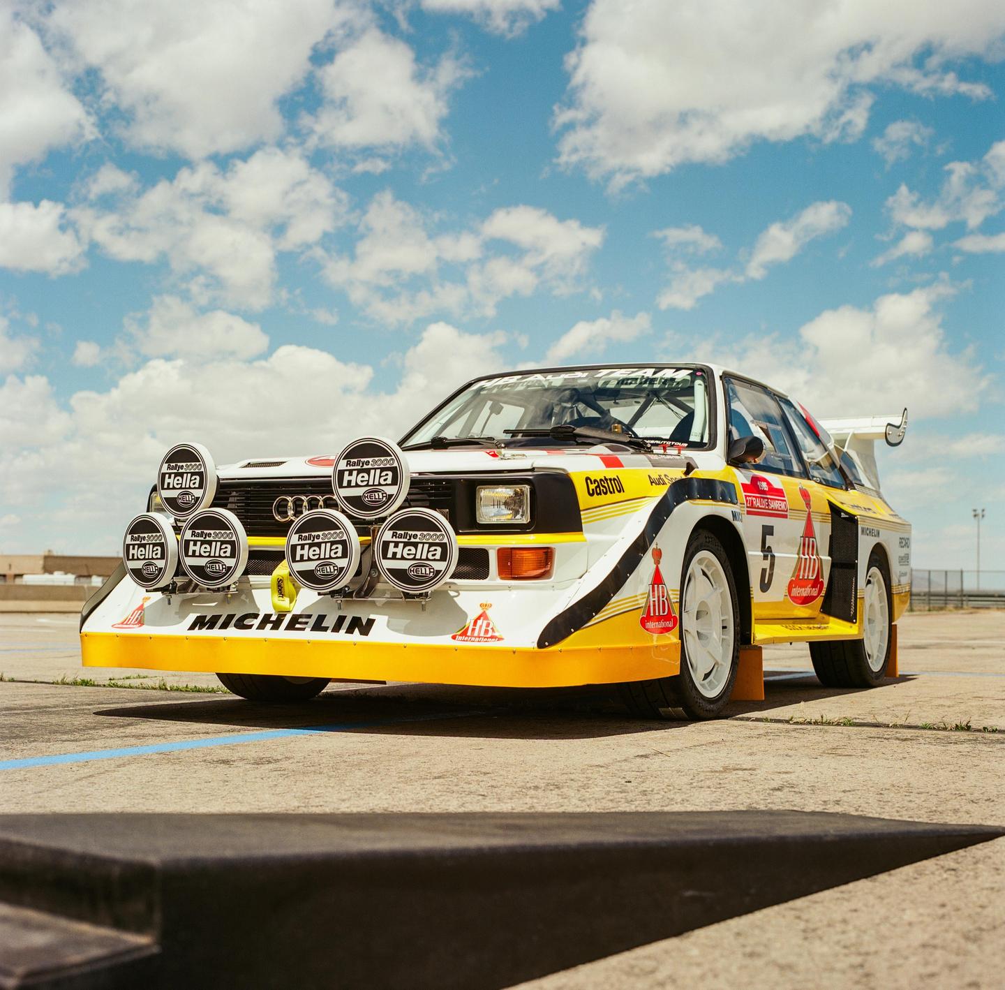 A white, yellow, and black rally car with many round lights on the front is parked on an asphalt surface under a blue sky with clouds.