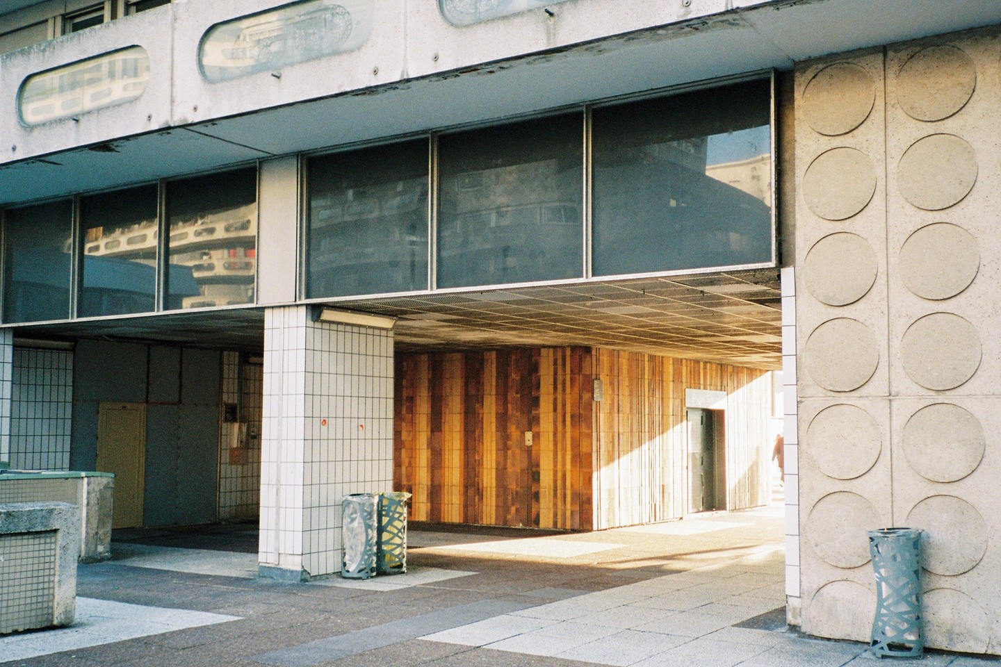 Sunlight illuminates a concrete and tiled underpass with a wooden wall and two trash cans.