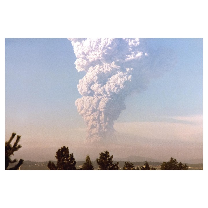 A large plume of smoke rises from a volcano above a distant town and trees.
