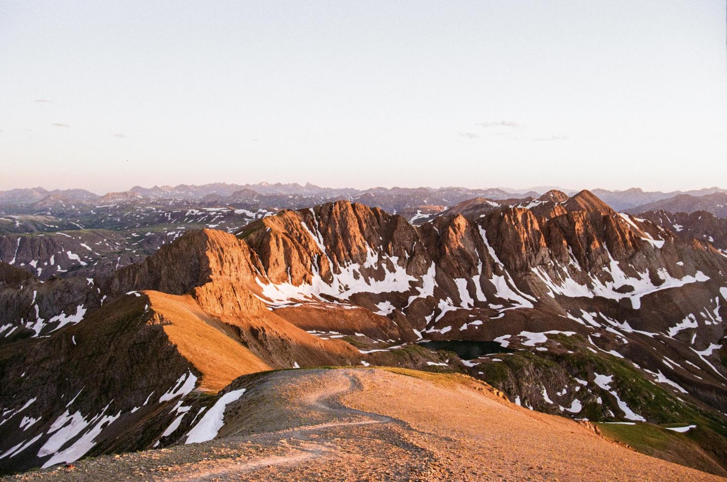 A winding trail leads up a mountain ridge with snow-capped peaks and a small lake visible in the distance under a clear sky.