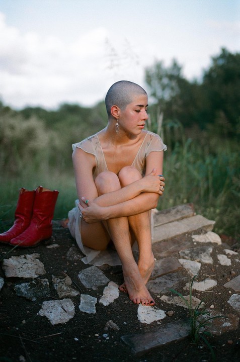 A woman with a shaved head sits barefoot on broken stones with red cowboy boots beside her.