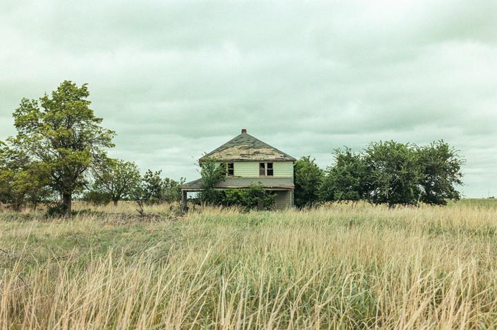 An abandoned house with a damaged roof stands in a field of tall grass under a cloudy sky.