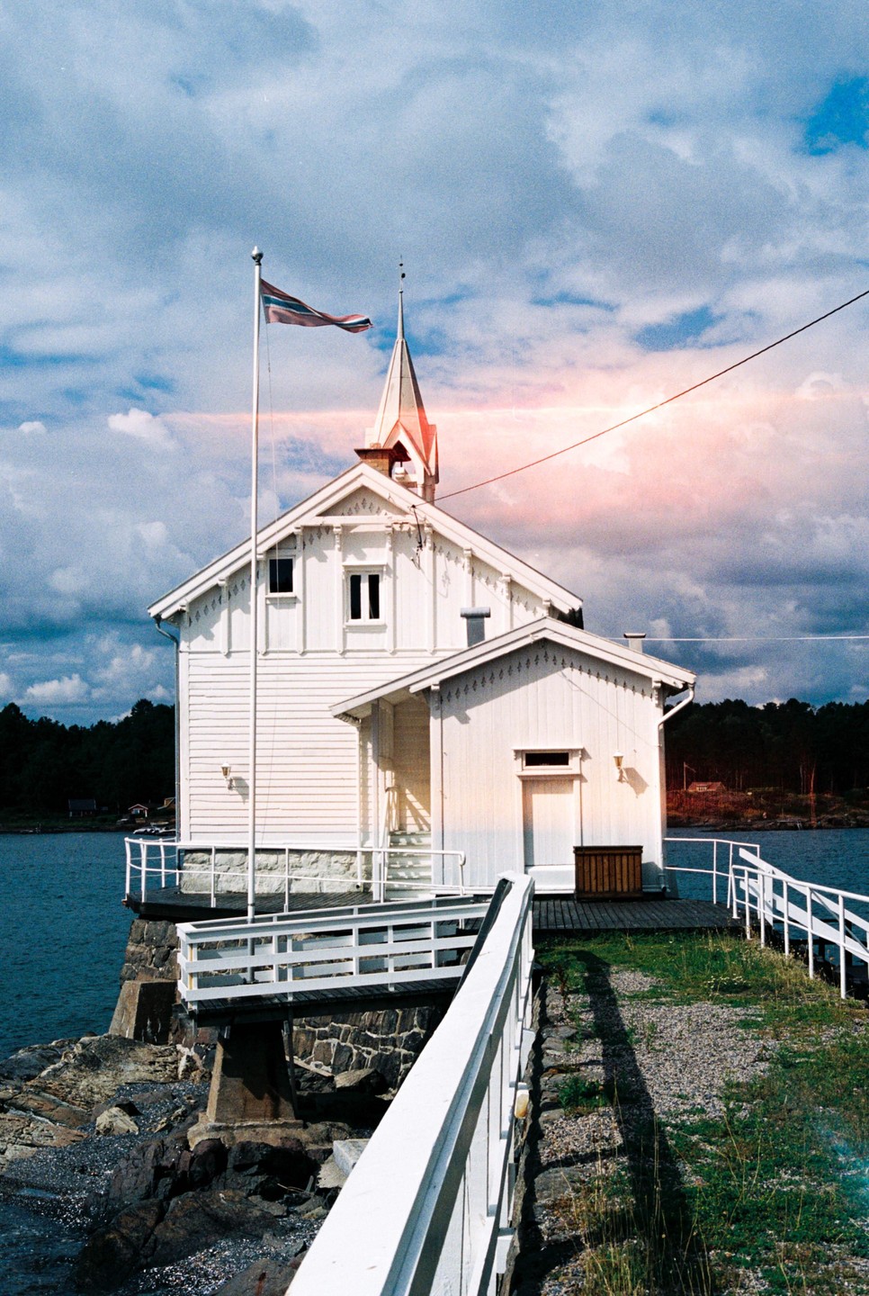 A white lighthouse with a spire and a flag stands on a rocky shore next to blue water under a cloudy sky.
