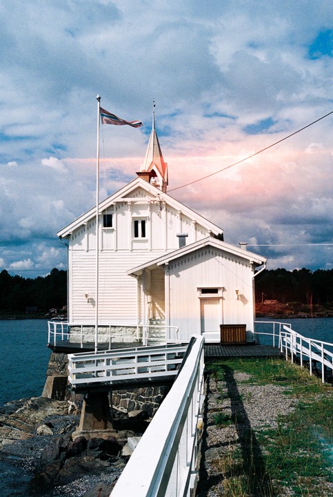 A white lighthouse with a spire and a flag stands on a rocky shore next to blue water under a cloudy sky.