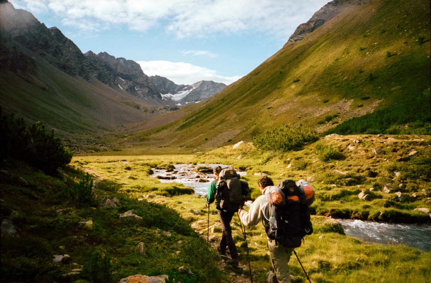 Two men with backpacks and hiking poles walk along a path next to a stream in a mountain valley.