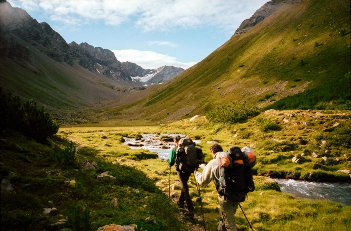 Two men with backpacks and hiking poles walk along a path next to a stream in a mountain valley.