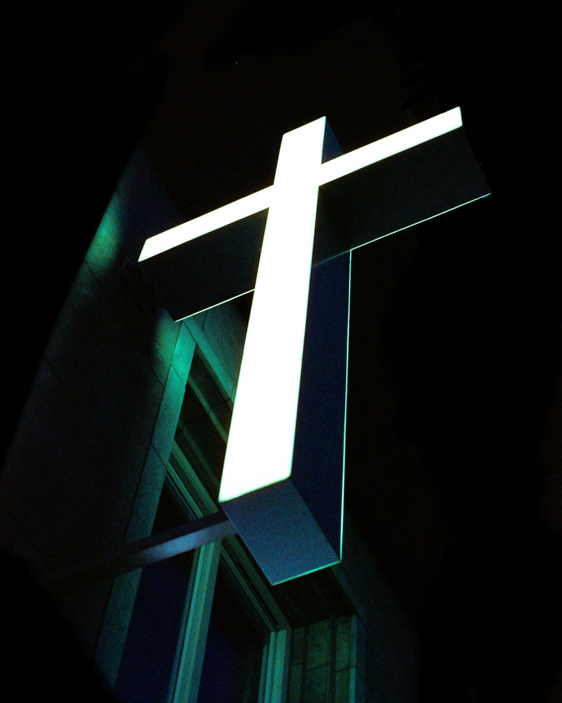 A bright white cross is illuminated against a dark background, with a building facade visible below it.
