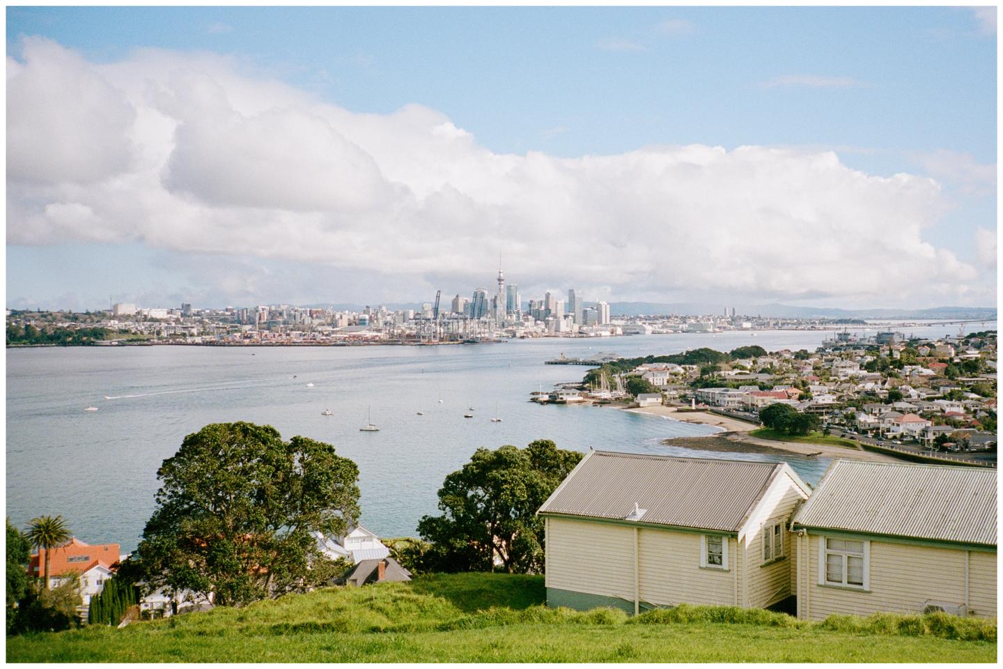 A panoramic view of Auckland, New Zealand, shows the city skyline across a body of water with boats, and houses in the foreground.