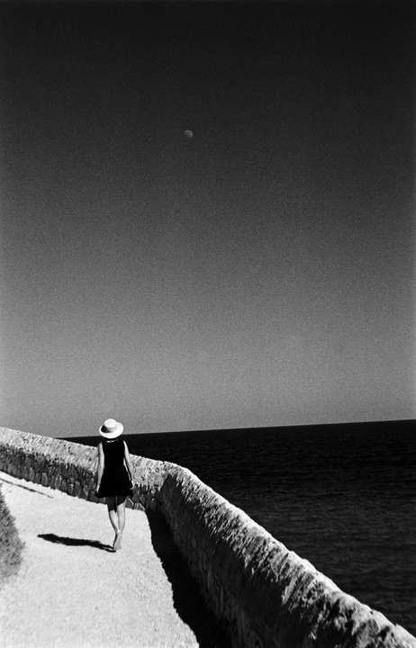 A woman in a hat and dress walks barefoot along a stone wall next to the ocean under a sky with a visible moon.