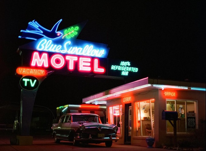 A vintage car is parked in front of the Blue Swallow Motel, illuminated by neon signs at night.