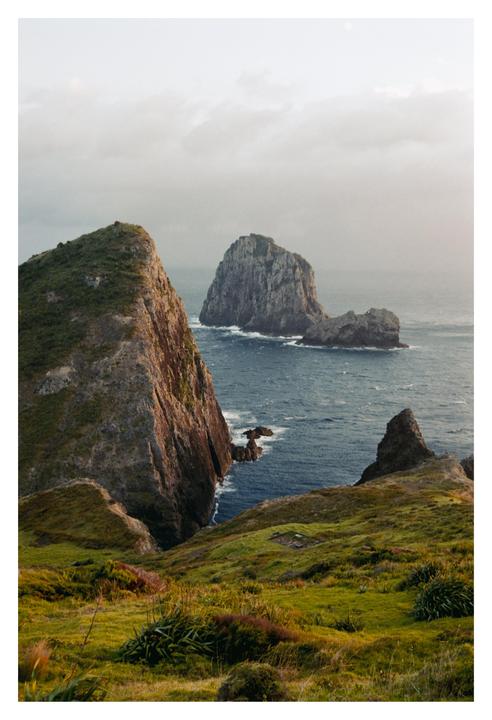A dramatic landscape shows a steep, grassy cliff overlooking the ocean with several rocky islands in the distance under a cloudy sky.