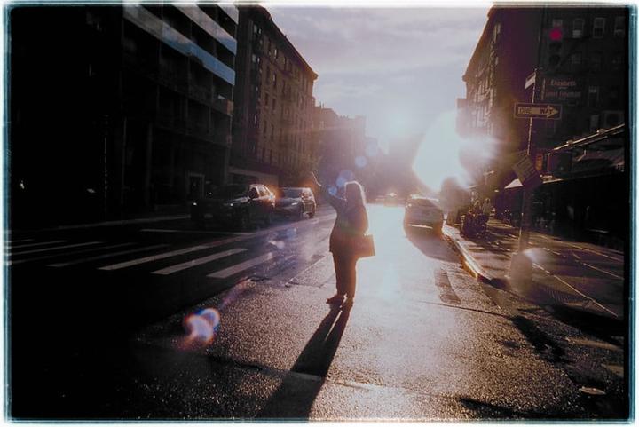 A woman stands on a wet street at sunset, with buildings and cars visible in the background.