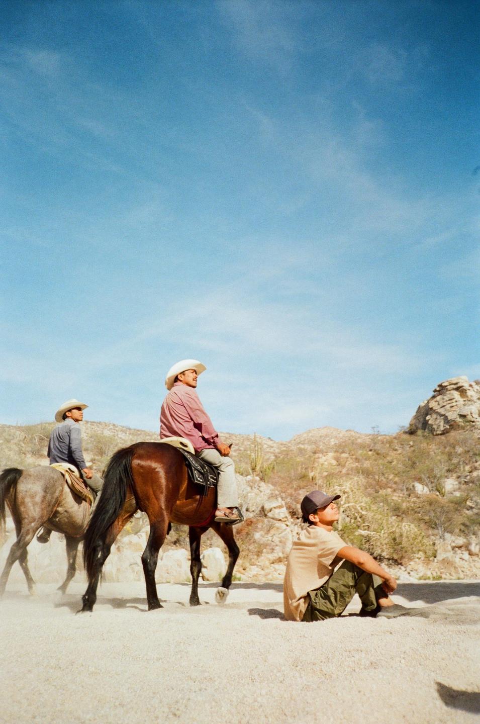 Two men on horseback and one man sitting on the ground are in a desert landscape under a clear sky.