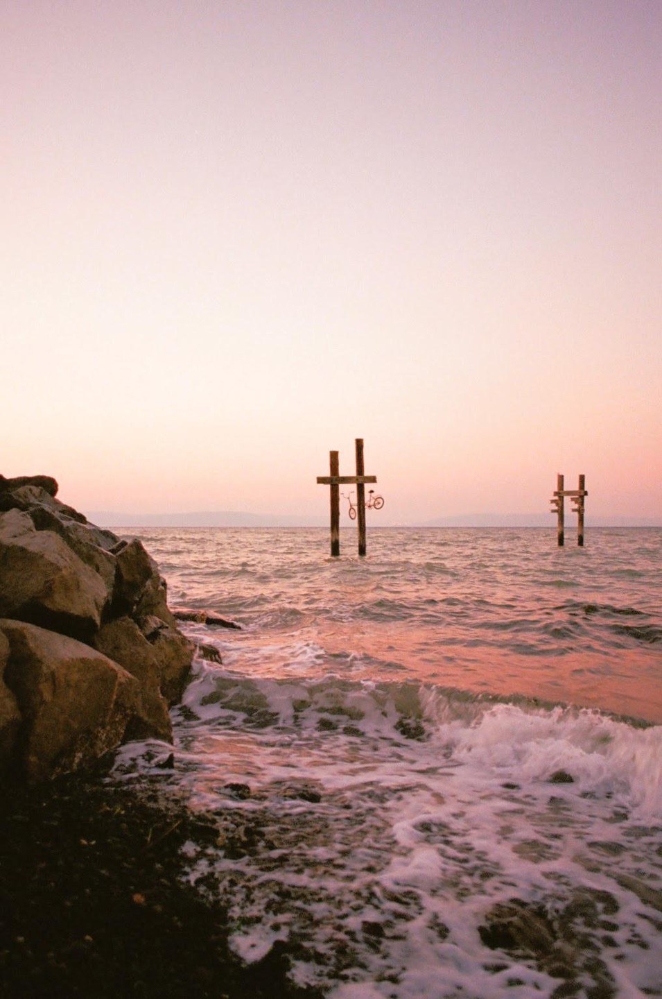 A bicycle hangs from a wooden structure in the water next to a rocky shore at sunset.