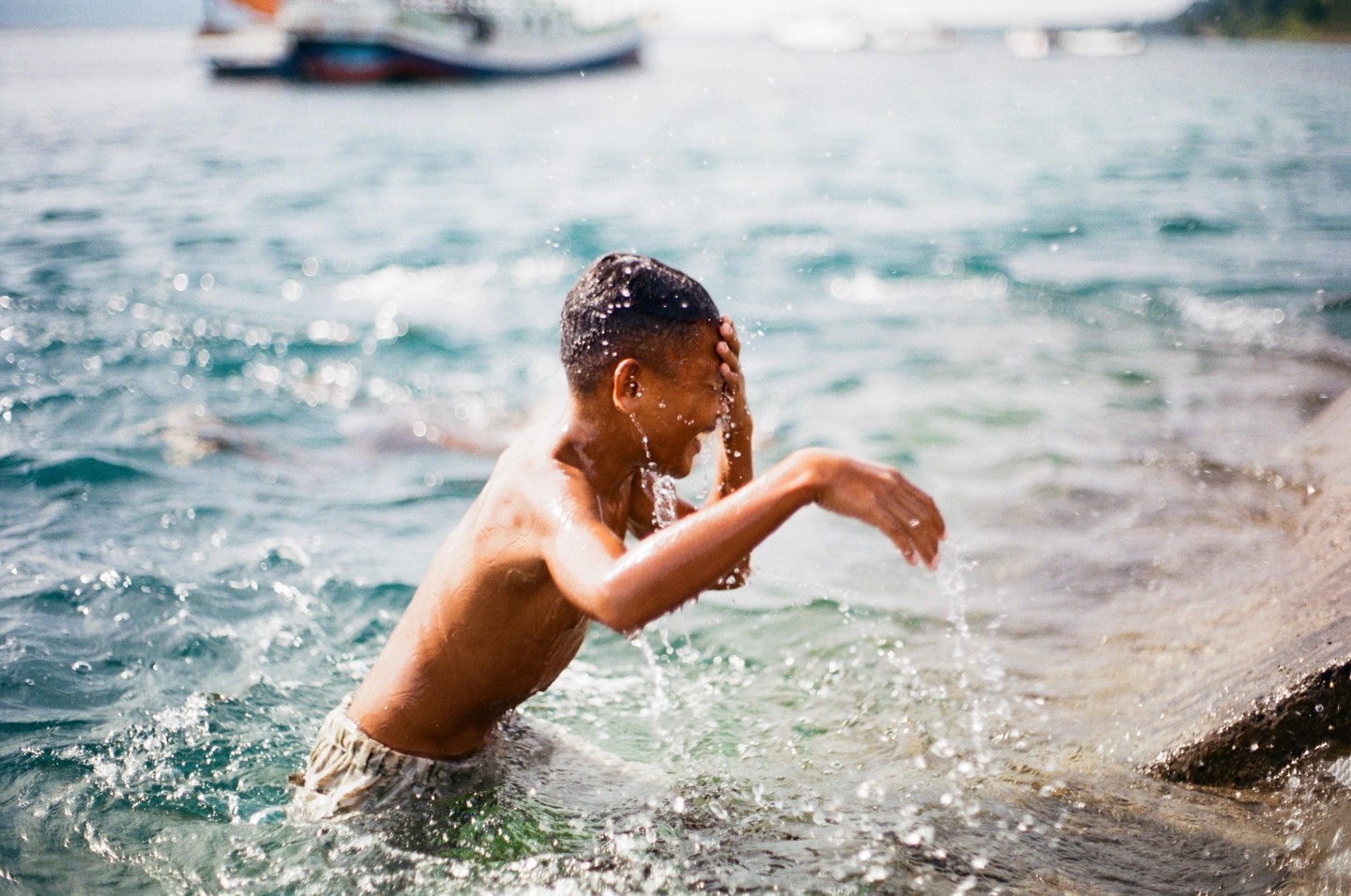 A boy splashes water on his face while standing in the ocean with a boat in the background.